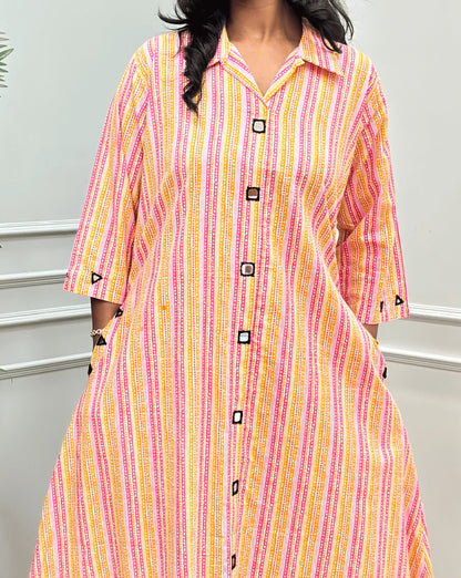 Close-up View of Multi Women's Ajrakh A-line Pure Cotton Long Kurti Featured with Collar V neck & 3/4th Quarter Sleeves with an Ajrakh Strip Print on abstract of Pink, White, Yellow, Orange, Real hand-mirror work on front & Sleeves, Along with 2 side pockets, standing on a round woven rug in front of a light gray paneled wall with a potted plant.