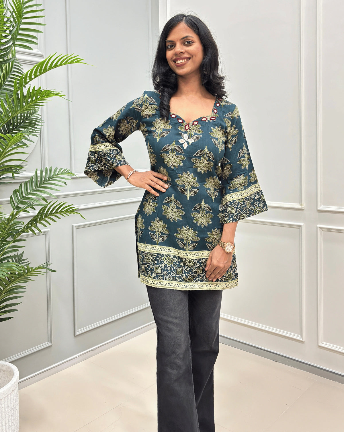 Green women's short kurti with three-quarter wide sleeves, ajrakh floral print , paired with black jeans , standing in front of a white paneled wall.