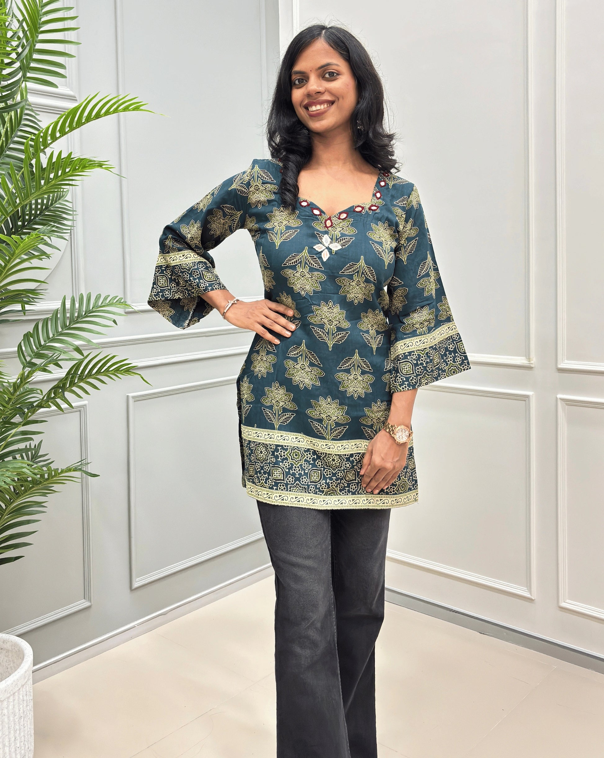Green women's short kurti with three-quarter wide sleeves, ajrakh floral print , paired with black jeans , standing in front of a white paneled wall.