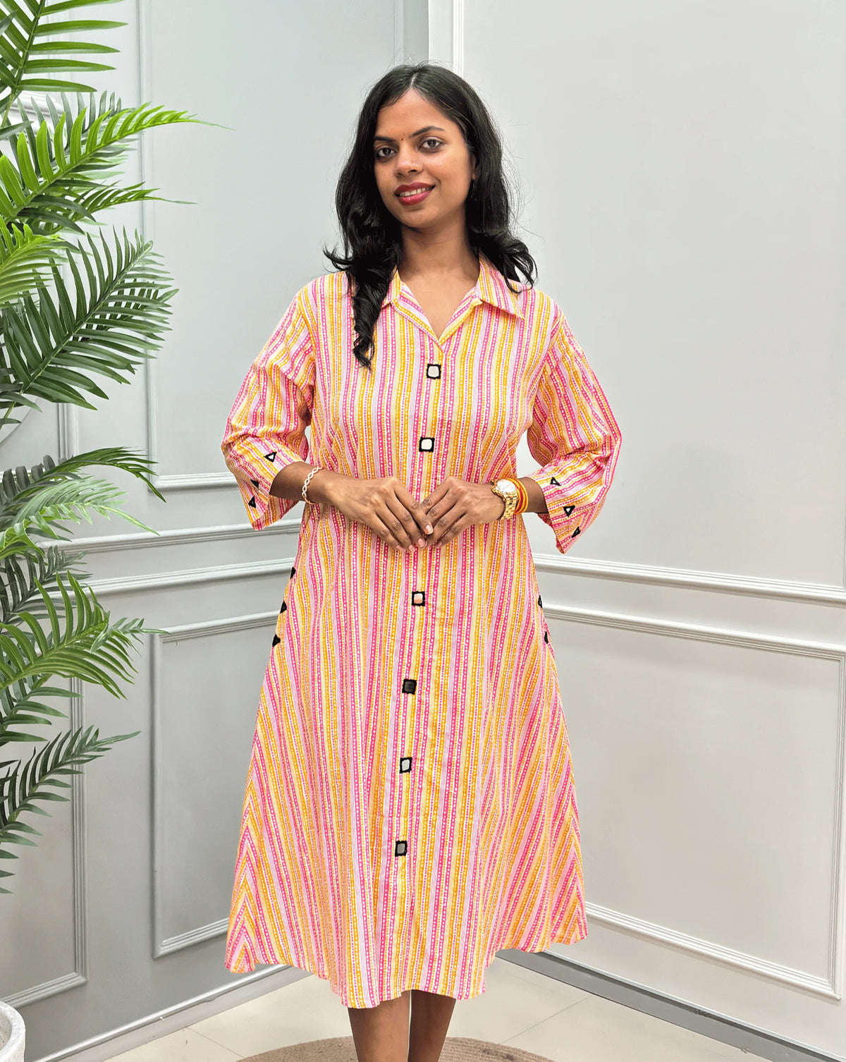 Front-View of Multi Women's Ajrakh A-line Pure Cotton Long Kurti with an Ajrakh Strip Print on abstract of Pink, White, Yellow, Orange, Real hand-mirror work on front & Sleeves, Along with 2 side pockets, standing on a round woven rug in front of a light gray paneled wall with a potted plant.