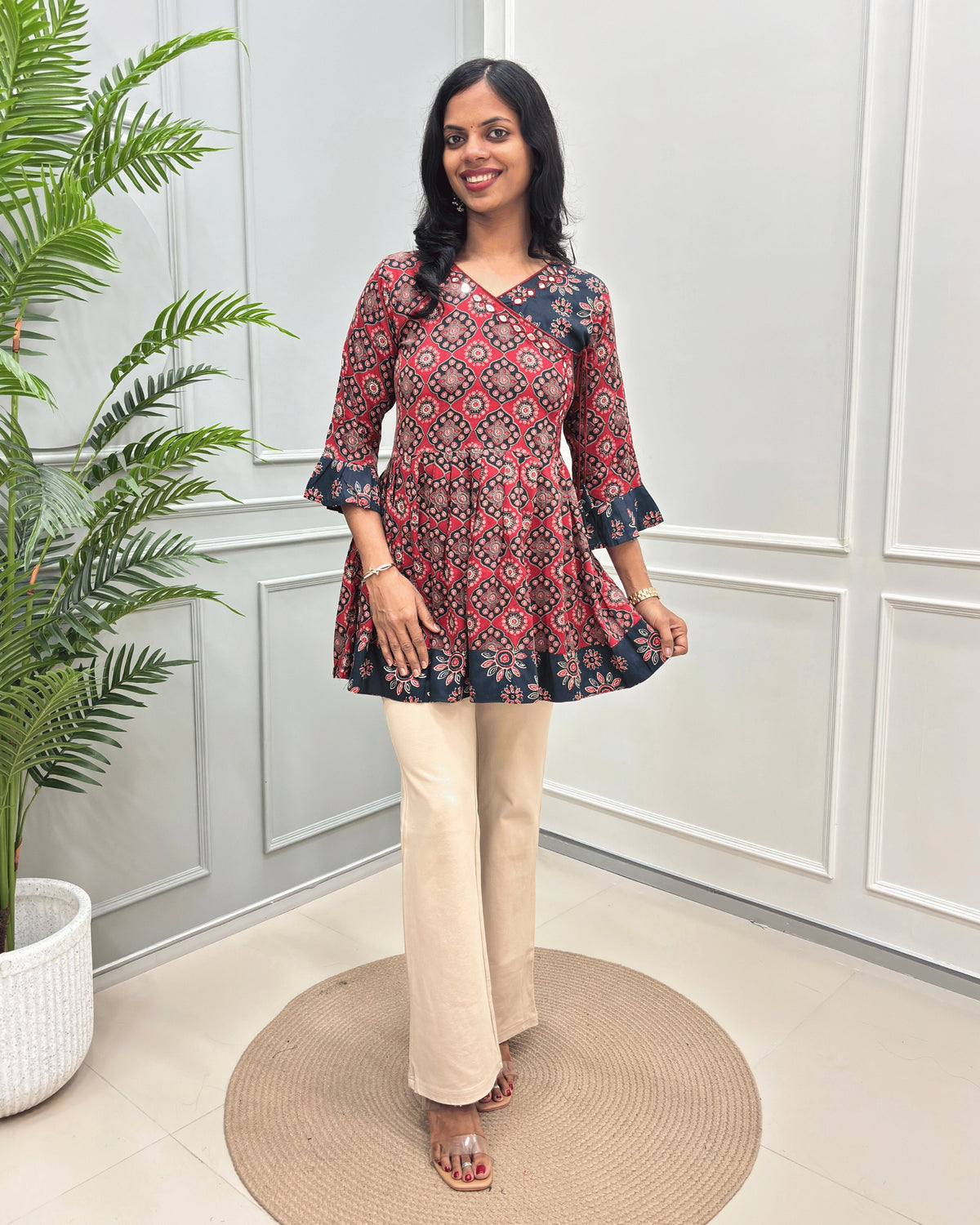 Red Women's Ajrakh Vegetable dyed Plant based Rayon Cotton with a Red, & blue geometric pattern on neck, sleeves cuffs & bottom  paired with beige wide-leg trousers, standing on a round woven mat in front of a white paneled wall.