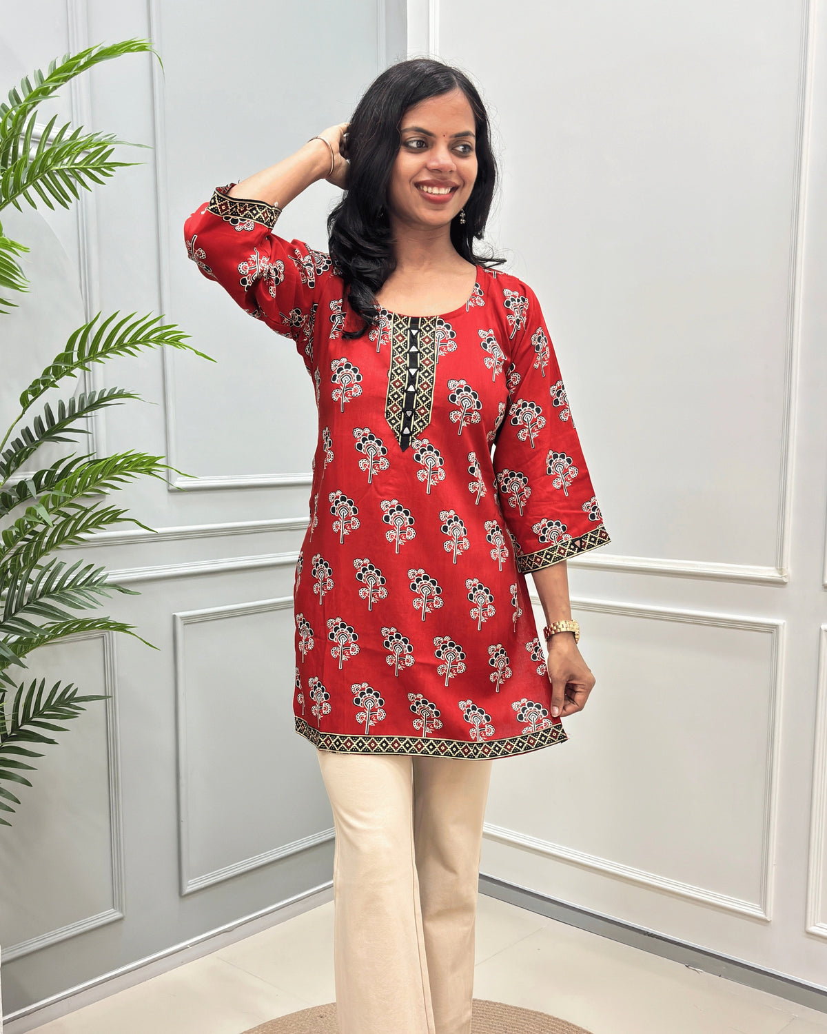 Front view of Red women's Ajrakh plant based cotton Short kurti with a colorful floral print, paired with beige pant, standing in front of a white paneled wall with a green plant on the left. 