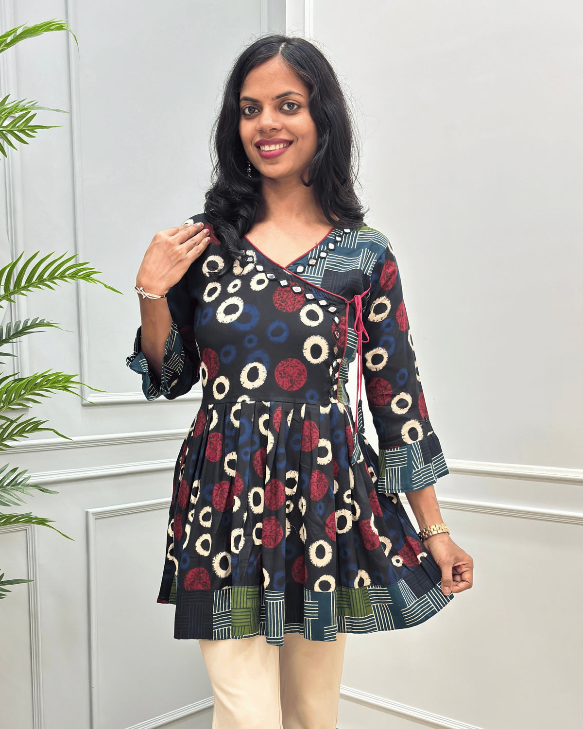 Close-up of Black women's Ajrakh plant based cotton Short kurti with a colorful geometric and circular print, Hand-Mirror work on V- neckline along with lace, paired with beige pants, standing in front of a white paneled wall with a green plant on the left.