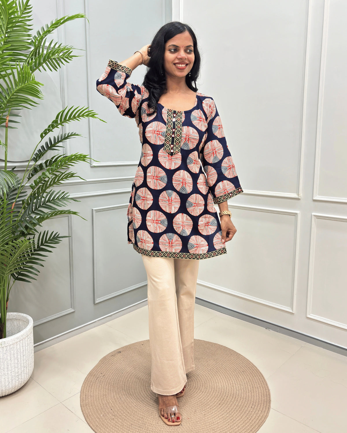 Navy-Blue women's Vegetable dyed plant based Cotton Short kurti with a colorful geometric circular print and beige border trim, paired with beige wide-leg trousers, standing on a round woven mat in front of a white paneled wall.