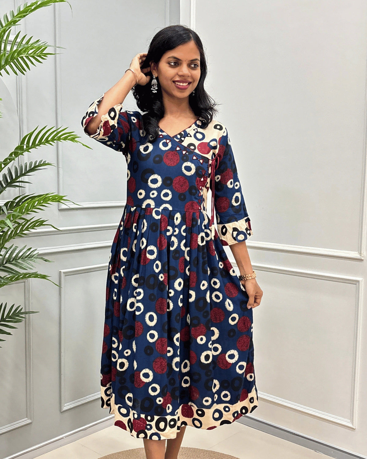 Front view of blue women's plant based rayon cotton long kurti with a red and cream circular dot pattern, along with frill sleeves and hand- mirror worked on V-neck standing in front of a white paneled wall with a green plant on the left.