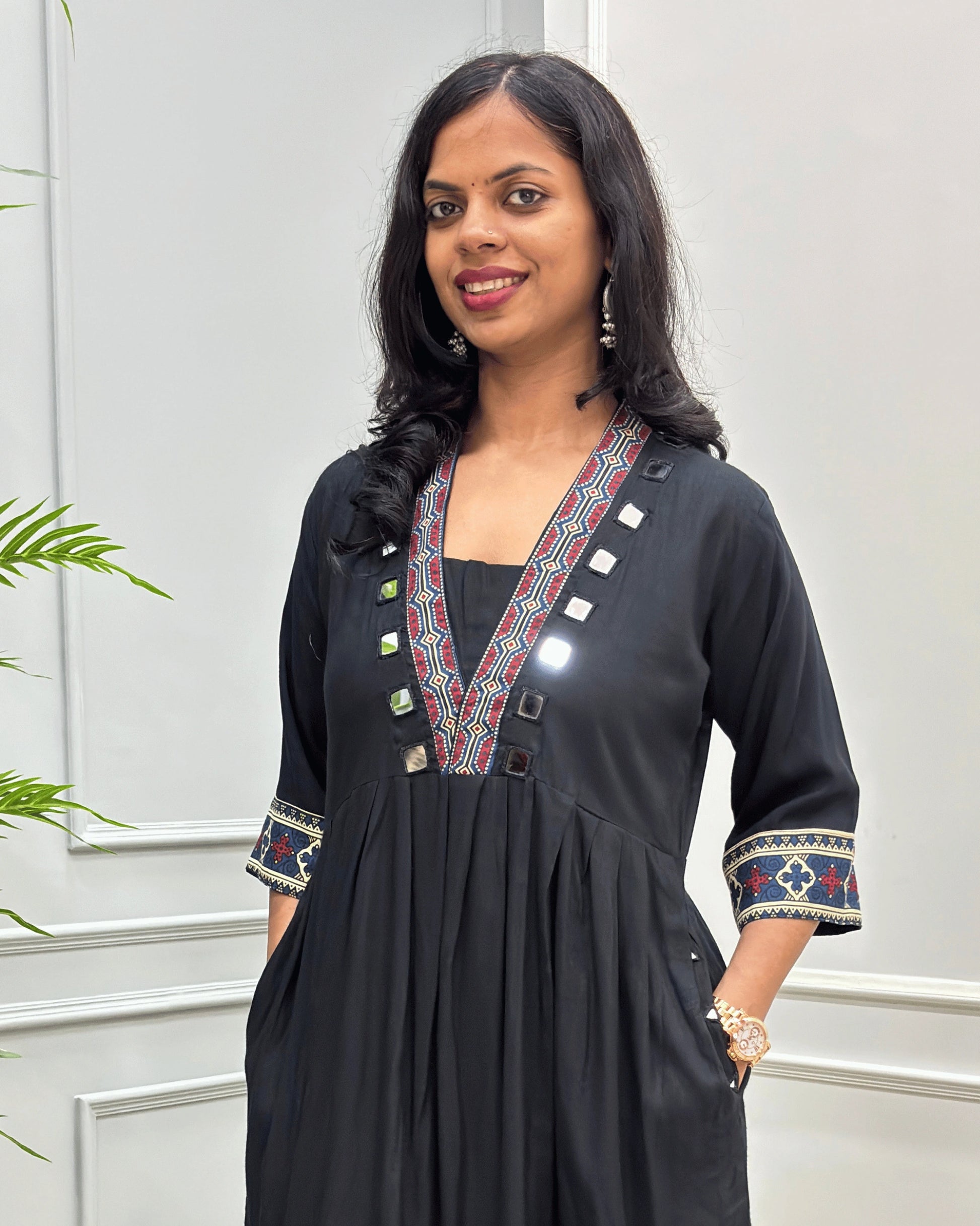 Close-up of Black women's plant based cotton long kurti with colorful embroidered trim on the neckline and cuffs,  mirror design on neck outline , 2 side pockets with hand mirror work,  standing against a light gray paneled wall.