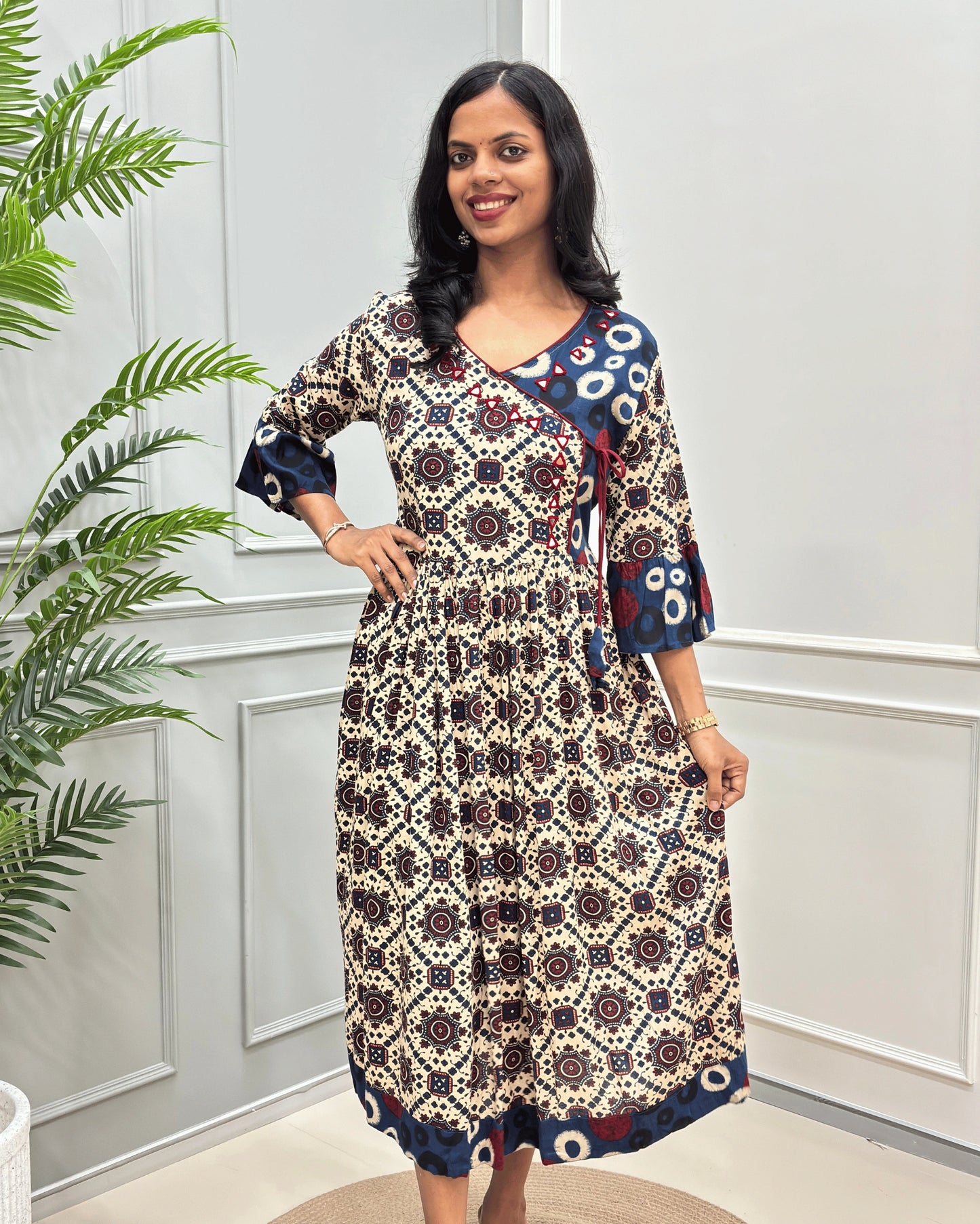 Front side of Beige woman's cotton long-kurti with three-quarter sleeve, with a geometric and floral mandala pattern in beige, navy, and red, standing in front of a white paneled wall with a green plant on the left.