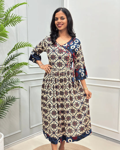 Front side of Beige woman's cotton long-kurti with three-quarter sleeve, with a geometric and floral mandala pattern in beige, navy, and red, standing in front of a white paneled wall with a green plant on the left.