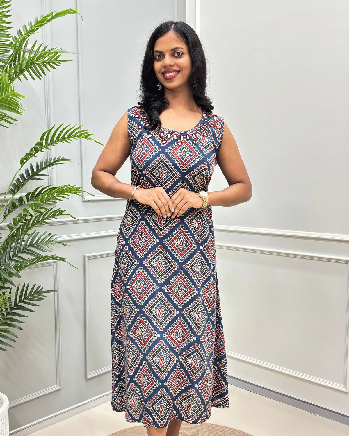 Front view of blue Cotton long sleeveless Kurti with a geometric diamond pattern in navy blue, red, and beige, standing in front of a white paneled wall with a green plant on the left.