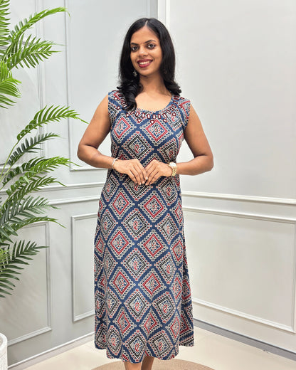 Front view of blue Cotton long sleeveless Kurti with a geometric diamond pattern in navy blue, red, and beige, standing in front of a white paneled wall with a green plant on the left.