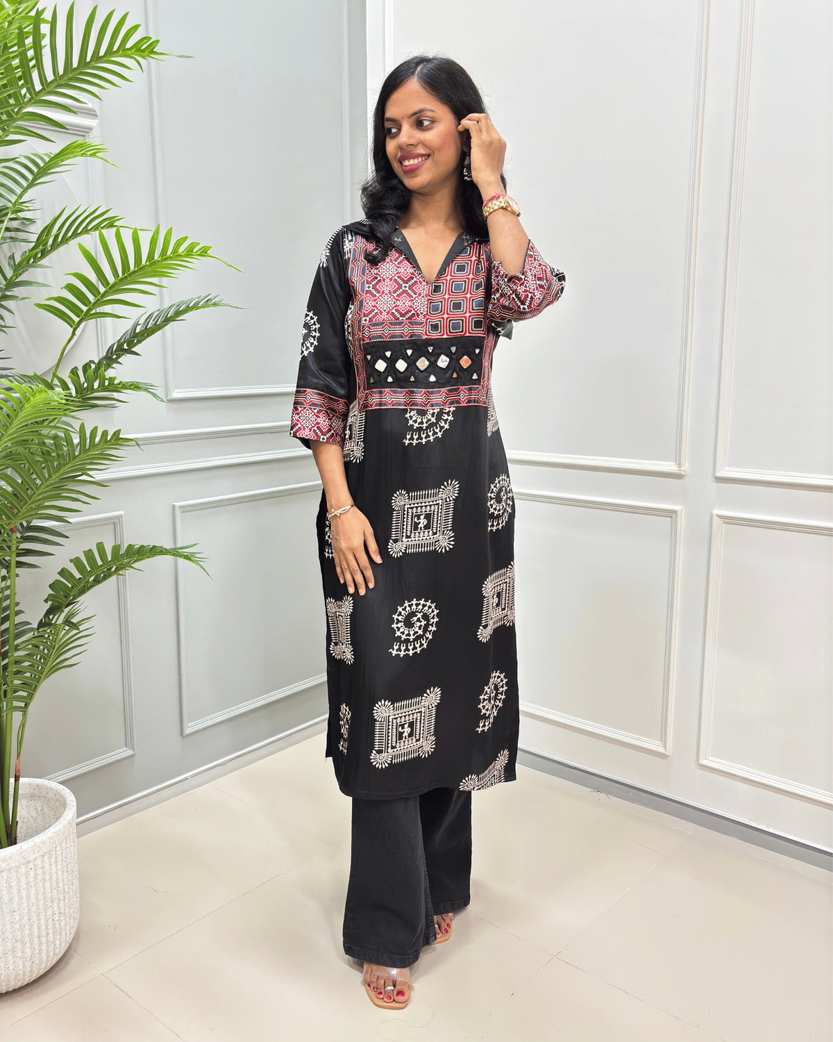 Black women's Pure Mashru Silk Long Kurti with red , Black & white geometric patterns on the yoke and sleeves, paired with black Flared pants, standing in a room with a potted plant.