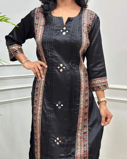 Close up of Black Women's Ajrakh Mashru Silk with multicolor Ajrakh print & Hand-Mirror embroidered Sleeves borders and Front panel standing indoors against a light gray paneled wall.