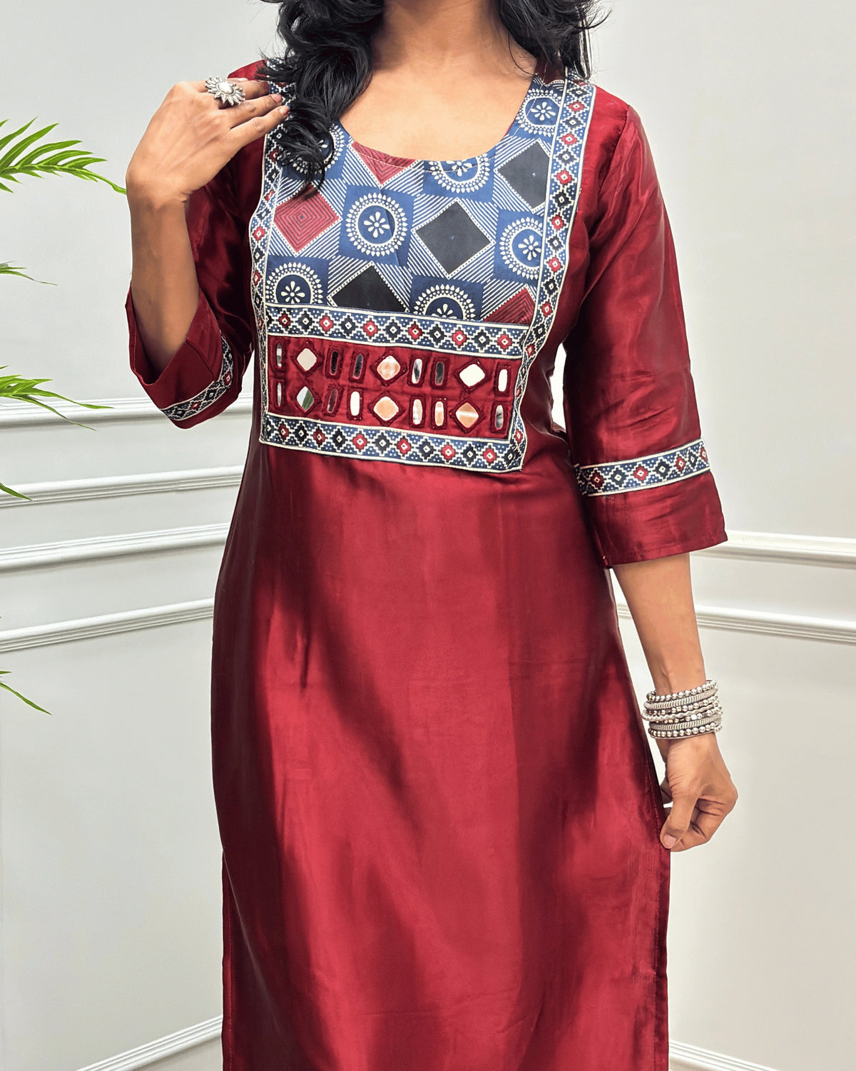 Close-up of Red Ajrakh Mashru Silk Long-kurti with a highly detailed geometric embroidered yoke in blue, red, and white, along with ajrakh print border on front & sleeves, posed against a light gray paneled wall.
