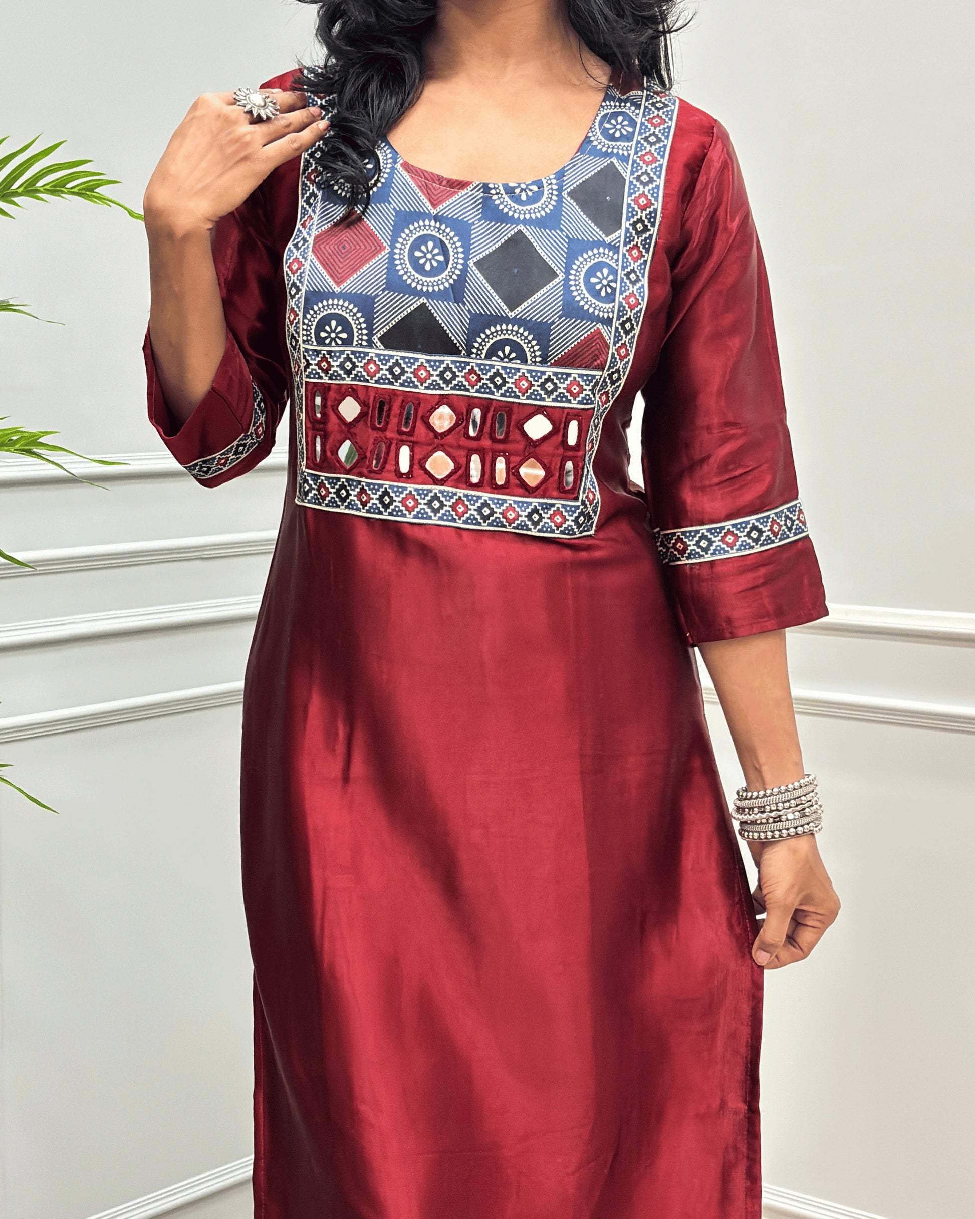 Close-up of Red Ajrakh Mashru Silk Long-kurti with a highly detailed geometric embroidered yoke in blue, red, and white, along with ajrakh print border on front & sleeves, posed against a light gray paneled wall.