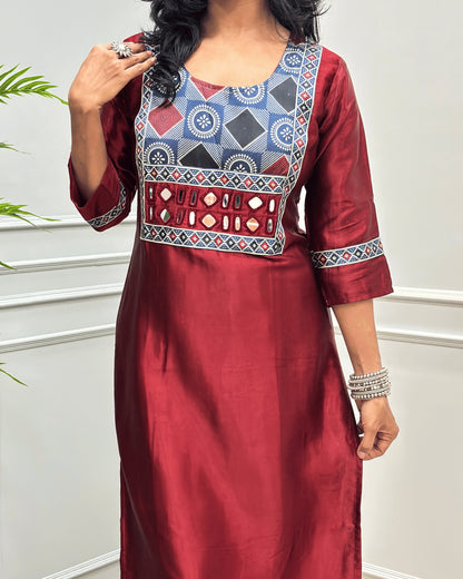 Close-up of Red Ajrakh Mashru Silk Long-kurti with a highly detailed geometric embroidered yoke in blue, red, and white, along with ajrakh print border on front & sleeves, posed against a light gray paneled wall.