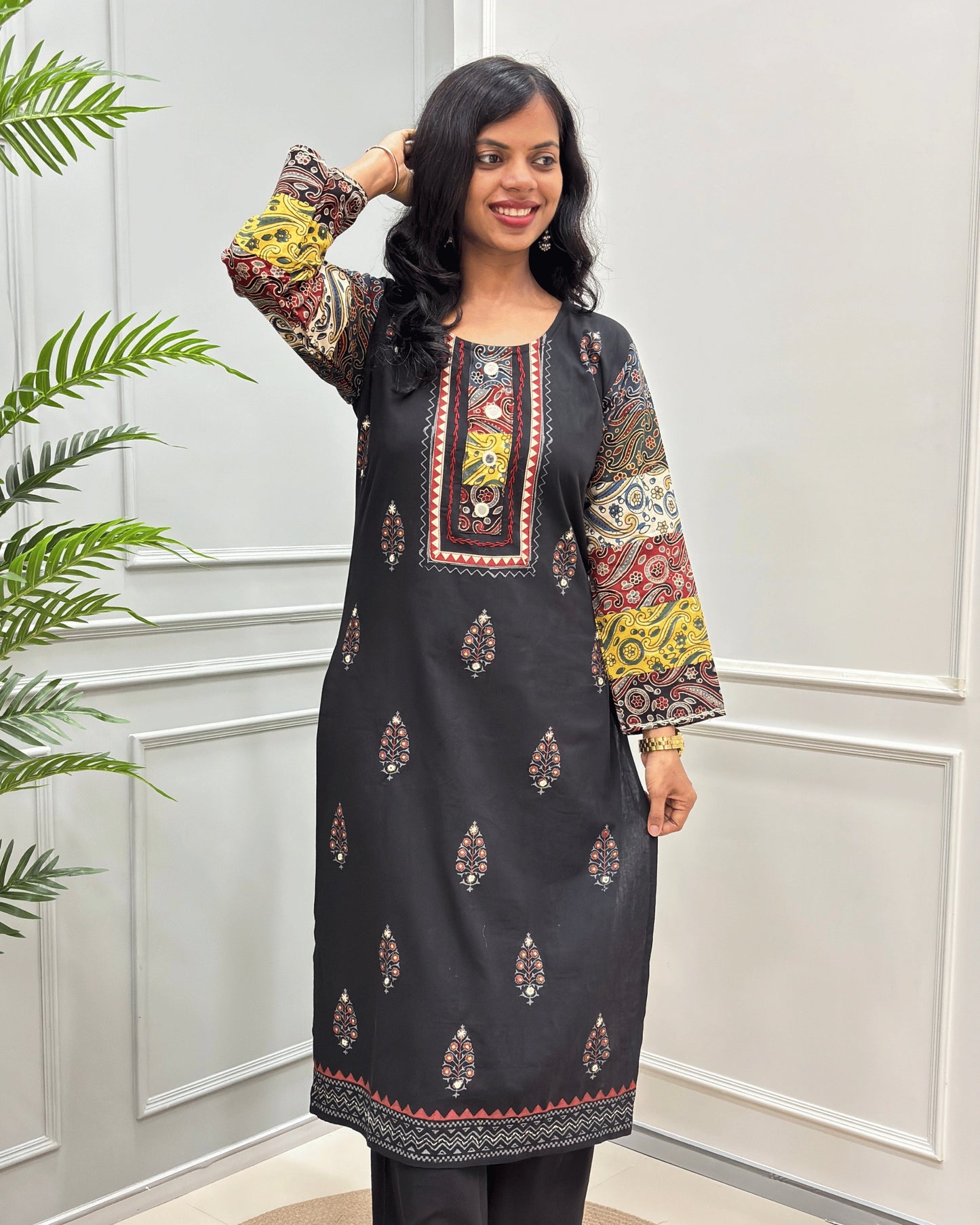 Front View of Black women's 2 Peice Ajrakh Cotton Co-Ord Set with multi-color printed sleeves & Front, Along with Leaf pattern, Beige Ajrakh print on bottom  and a matching Black Plazzo , standing in front of a white paneled wall with a green plant on the left.