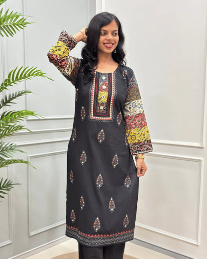 Front View of Black women's 2 Peice Ajrakh Cotton Co-Ord Set with multi-color printed sleeves & Front, Along with Leaf pattern, Beige Ajrakh print on bottom  and a matching Black Plazzo , standing in front of a white paneled wall with a green plant on the left.