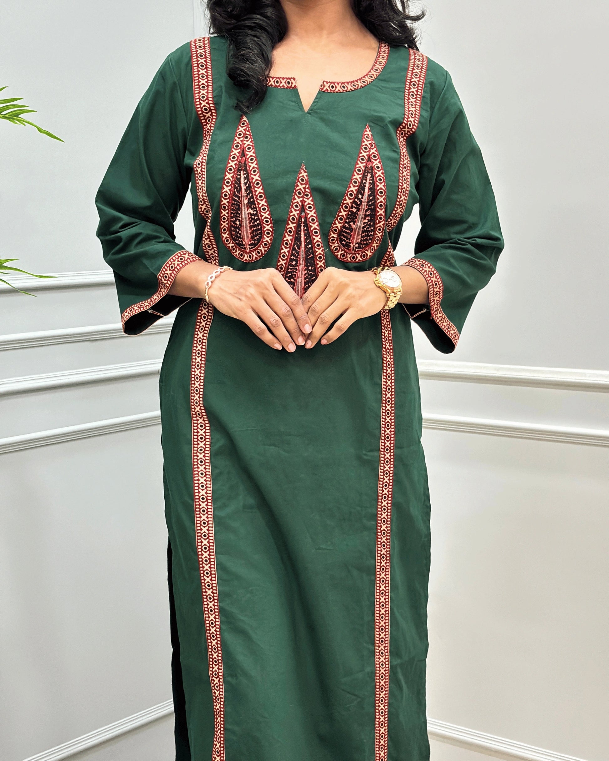 Close-up of Green Women's Hand-block  Ajrakh Pure Cotton Long-kurti with three-quarter sleeve featured with intricate Beige , Black and Red embroidered details on the neckline, cuffs, and front  panel, Along with Palm Leaf set against a light gray wall with a plant in the background.