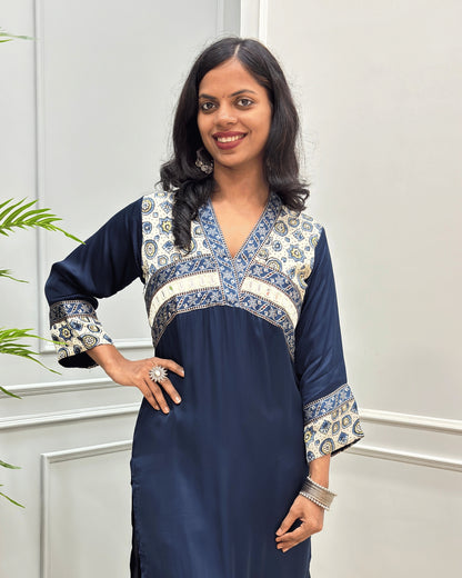 Close-up of Navy-Blue Ajrakh Model silk fabric with three-quarter sleeves with intricate blue and white patterned embroidery on the V-neckline, chest panel, and cuffs, standing in a light-colored room. 
