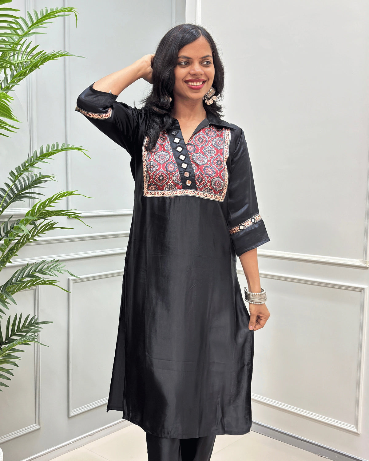 Front view of Black women's Ajrakh Mashru Silk with three-quarter Sleeves Featured with detailed multicolor embroidered yoke and patterned sleeve cuffs with Badam Buta print , standing in front of a white paneled wall with a green plant on the left.
