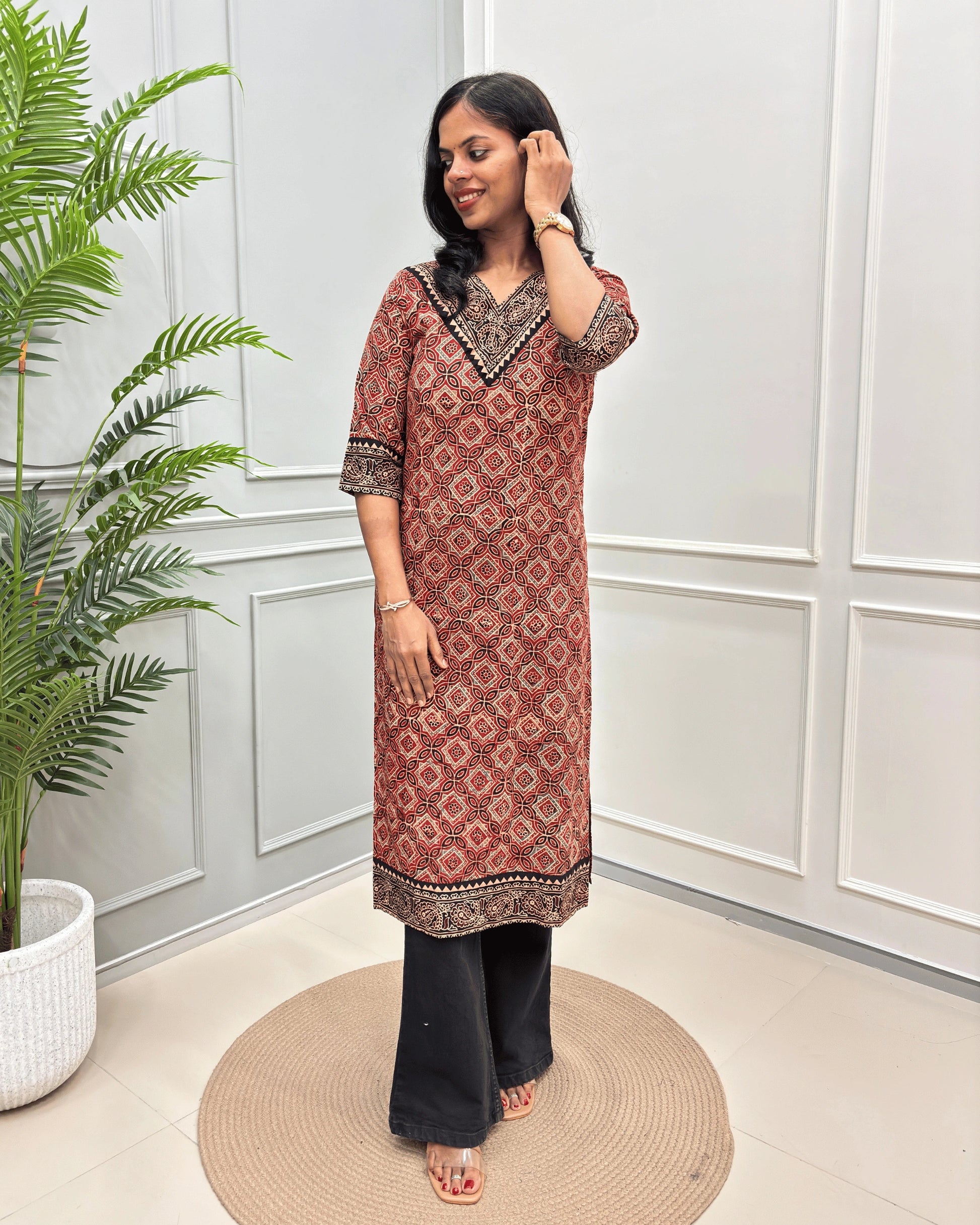 Red Women's Ajrakh Hand-block Pure Cotton Long-Kurti with Black and Beige geometric patterns, paired with Black Flared pants, standing on a round woven mat in front of a white paneled wall with a green potted plant.