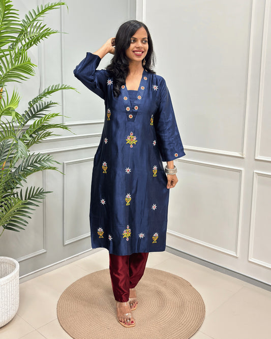 Navy Blue women's Pure Mashru Silk Long Kurti with yellow and white floral embroidery,& Ahir work on front & Sleeves cuffs , paired with maroon pants, standing on a round woven mat in front of a white paneled wall with a green plant to the side.