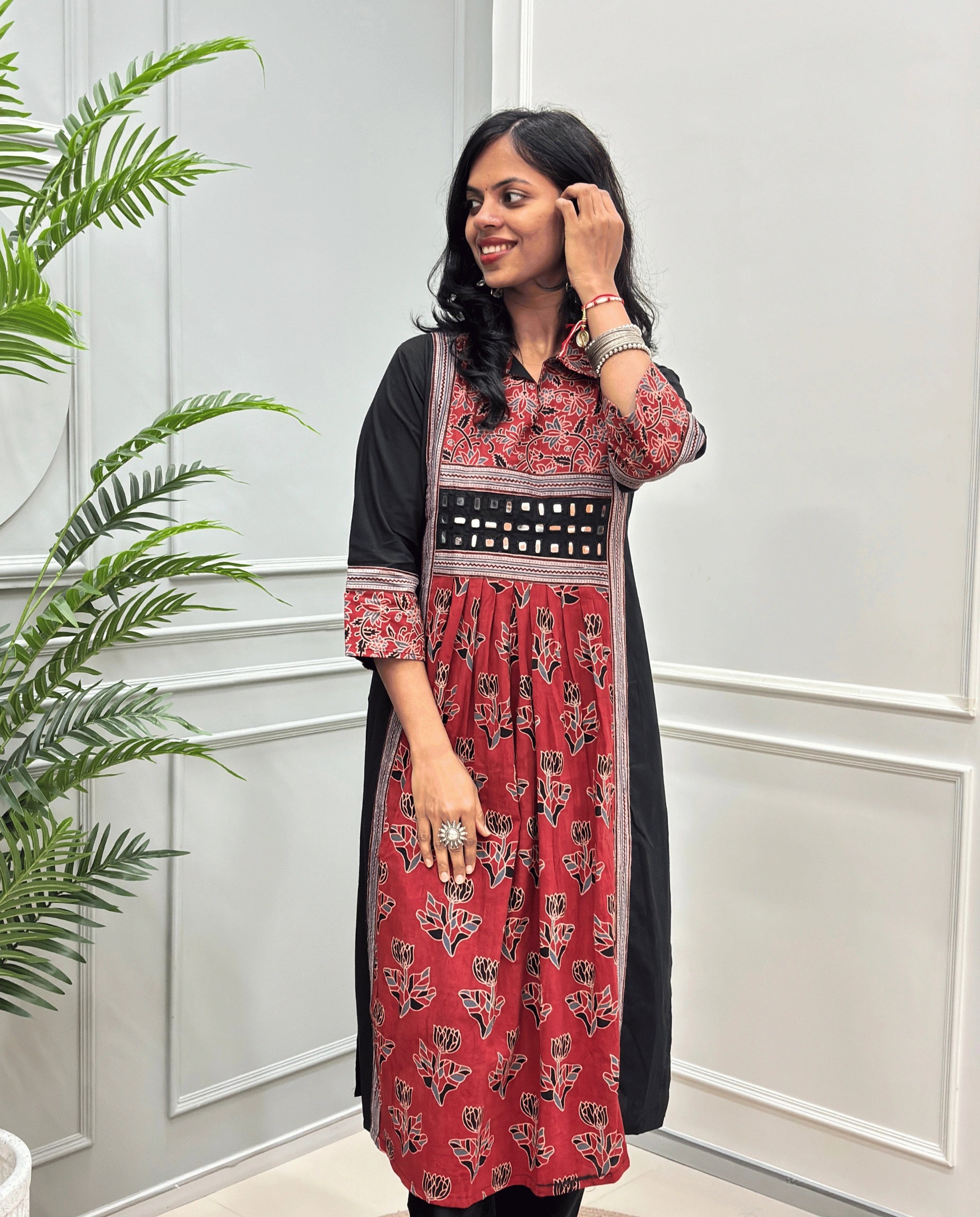 Front view of Red women's Ajrakh plant based cotton Long-kurti featured with a Black outer kurta and a Red printed inner kurta with floral motifs on front & sleeves , set against a white paneled wall with a green plant on the left.