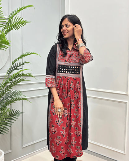 Front view of Red women's Ajrakh plant based cotton Long-kurti featured with a Black outer kurta and a Red printed inner kurta with floral motifs on front & sleeves , set against a white paneled wall with a green plant on the left.