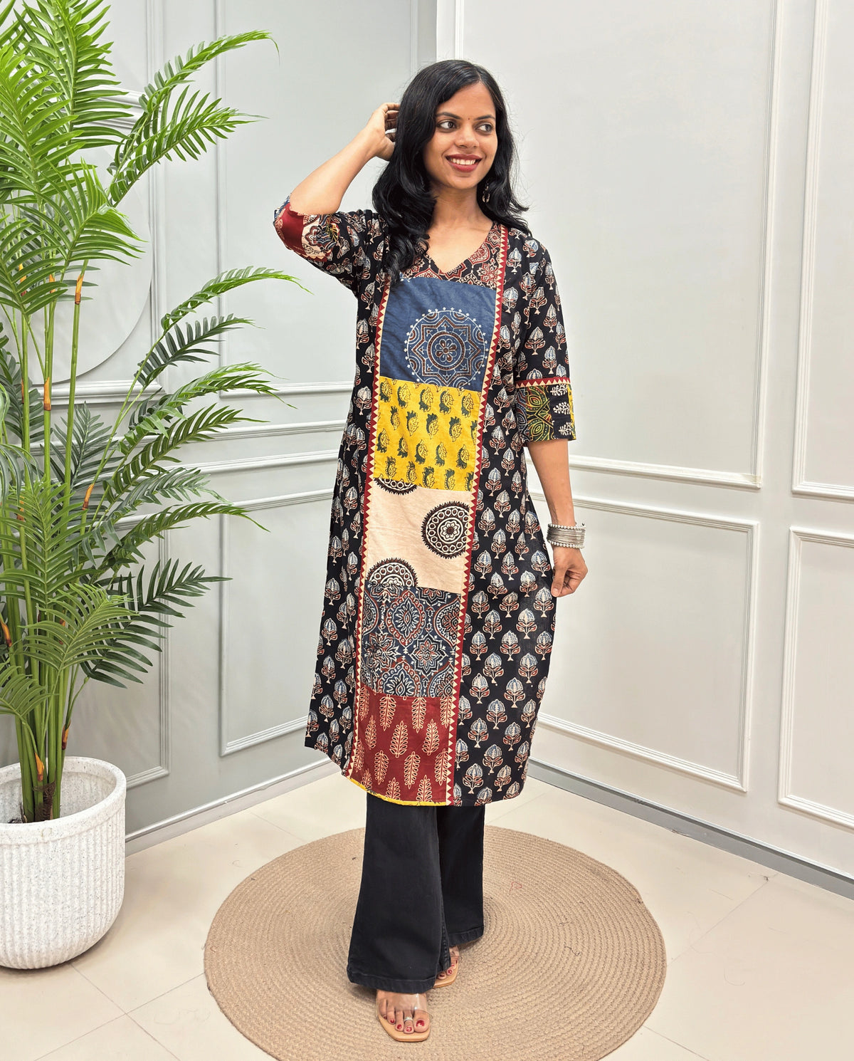 Black women's Hand-block Ajrakh  pure cotton Long Kurti with three-quarter sleeves, Ajrakh print on front and sleeves and black wide-leg pants, standing on a round woven mat in front of a white paneled wall next to a potted palm.