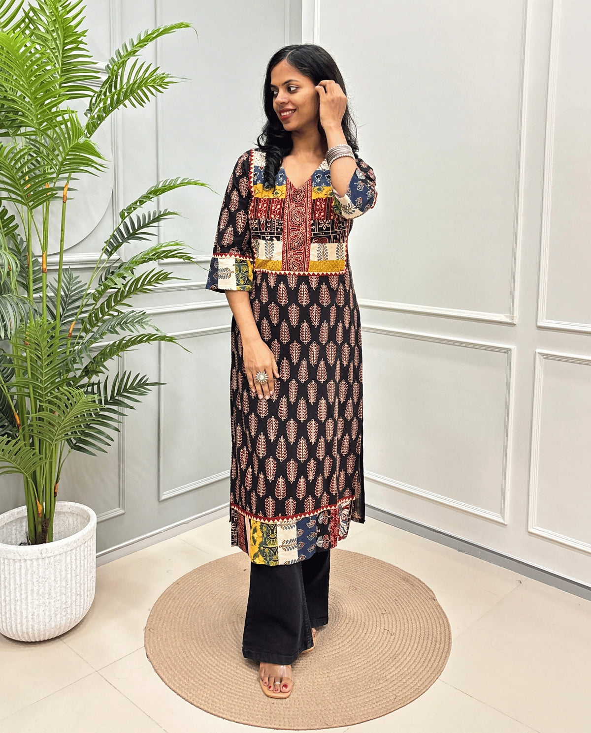 Black Women's Hand-block Ajrakh Pure cotton Long-Kurti with multicolor patchwork-style yoke, Cuffs and hem panels, paired with black Flared pants, standing indoors against a white paneled wall next to a potted plant.