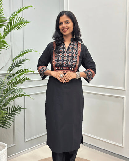 Front view of Black Women's Hand-block Ajrakh Pure Cotton Long kurti with intricate green geometric print on the chest and sleeve cuffs, hand-mirror work on front , paired with black pants , standing on a round woven mat in front of a white paneled wall with a potted plant.