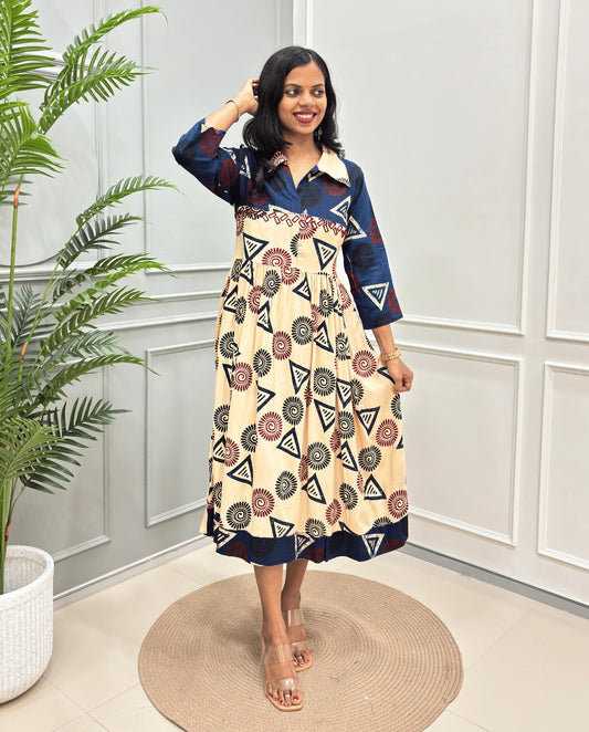 Beige Women's Ajrakh Vegetable dyed plant based Cotton Long-Kurti with a navy blue neck and cuffs, and a cream-colored dress featuring a geometric pattern of triangles and spiral in red, black, and blue, standing on a round woven rug in front of a white paneled wall with a green potted plant on the left.