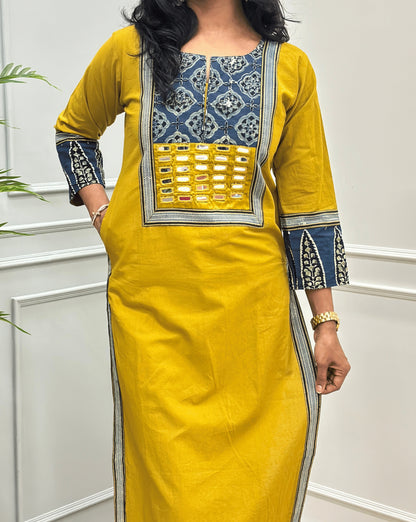 Close-up of Black Women's Hand-block Ajrakh Pure Cotton Long Kurti Featured with Three-quarter Sleeves & Round neck with V-shape cut with a Blue base and abstract Blue, Black & Beige geometric embroidery on the chest and sleeve cuffs, Hand-mirror work on Front, Ajrakh print border on Sides & Front, with Side pockets paired with black Flared pants, standing in a decorated room with a potted plant