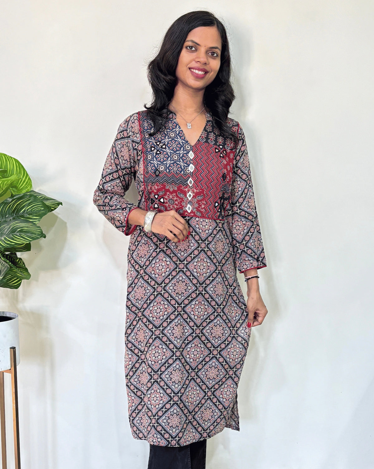 Front view of Black Women's Ajrakh Vegetable Dyed plant based cotton long kurti with black, and red patterns, Hand-mirror work on front & Sleeves paired with black flared pants, standing in a room with a white wall and a potted plant.