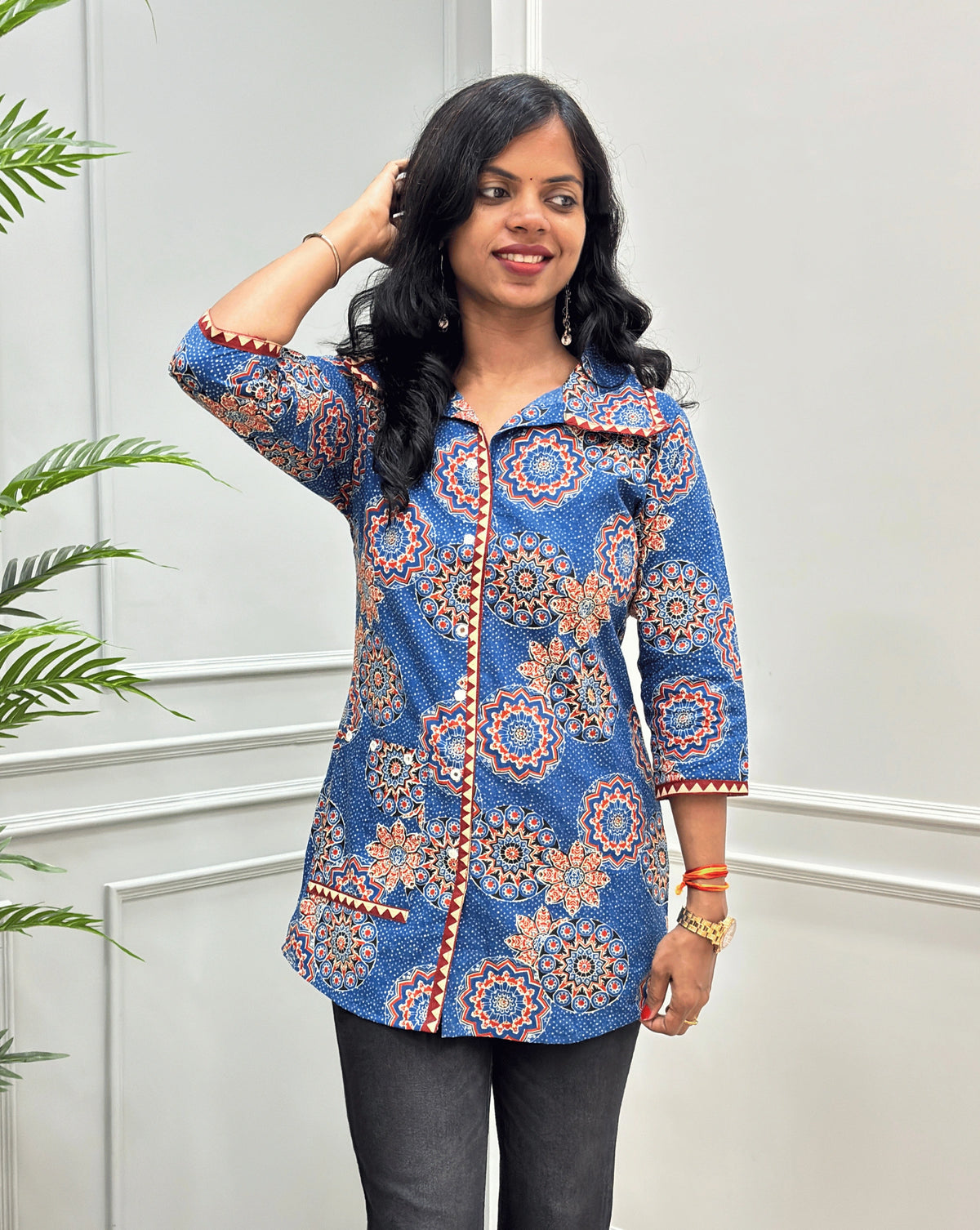 Front View of Blue women's Ajrakh pure cotton Shirt with a Blue base and Multi color floral print, Hand-mirror work on front button style, Sleeves cuffs & pocket, paired with black flared pants. Standing on a round woven rug against a light paneled wall with a potted plant at left.