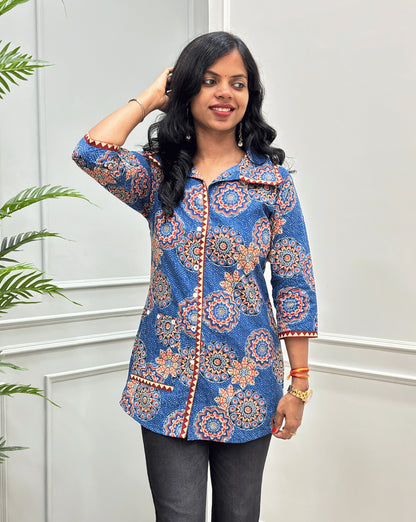 Front View of Blue women's Ajrakh pure cotton Shirt with a Blue base and Multi color floral print, Hand-mirror work on front button style, Sleeves cuffs & pocket, paired with black flared pants. Standing on a round woven rug against a light paneled wall with a potted plant at left.