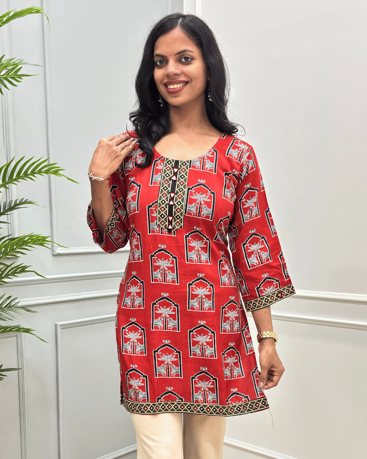 Close-up of Red women's Ajrakh plant based cotton short kurti with a traditional Indian arch pattern in black, white, and gold, hand-mirror work on front near Round neck , Ajrakh print on front, bottomm sleeves cuffs , paired with beige flared pant , standing against a white paneled wall with a green plant on the left.