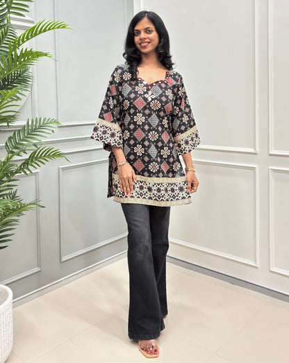 Black Women's cotton Short Kurti printed with beige and red geometric patterns, paired with black flare jeans, standing in a brightly lit room with white paneled walls and a potted plant on the left.