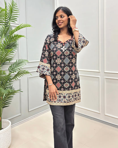 Women wearing a Black short kurti with intricate multi-color geometric & floral print, Ajrakh print on border  paired with black jeans.