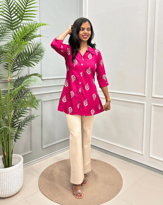 Deep Pink Women's Ajrakh A-line Pure Cotton Short Kurti with a White Ajrakh Leaf Print, paired with beige Flared trousers, standing on a round woven mat in front of a white paneled wall with a potted plant.