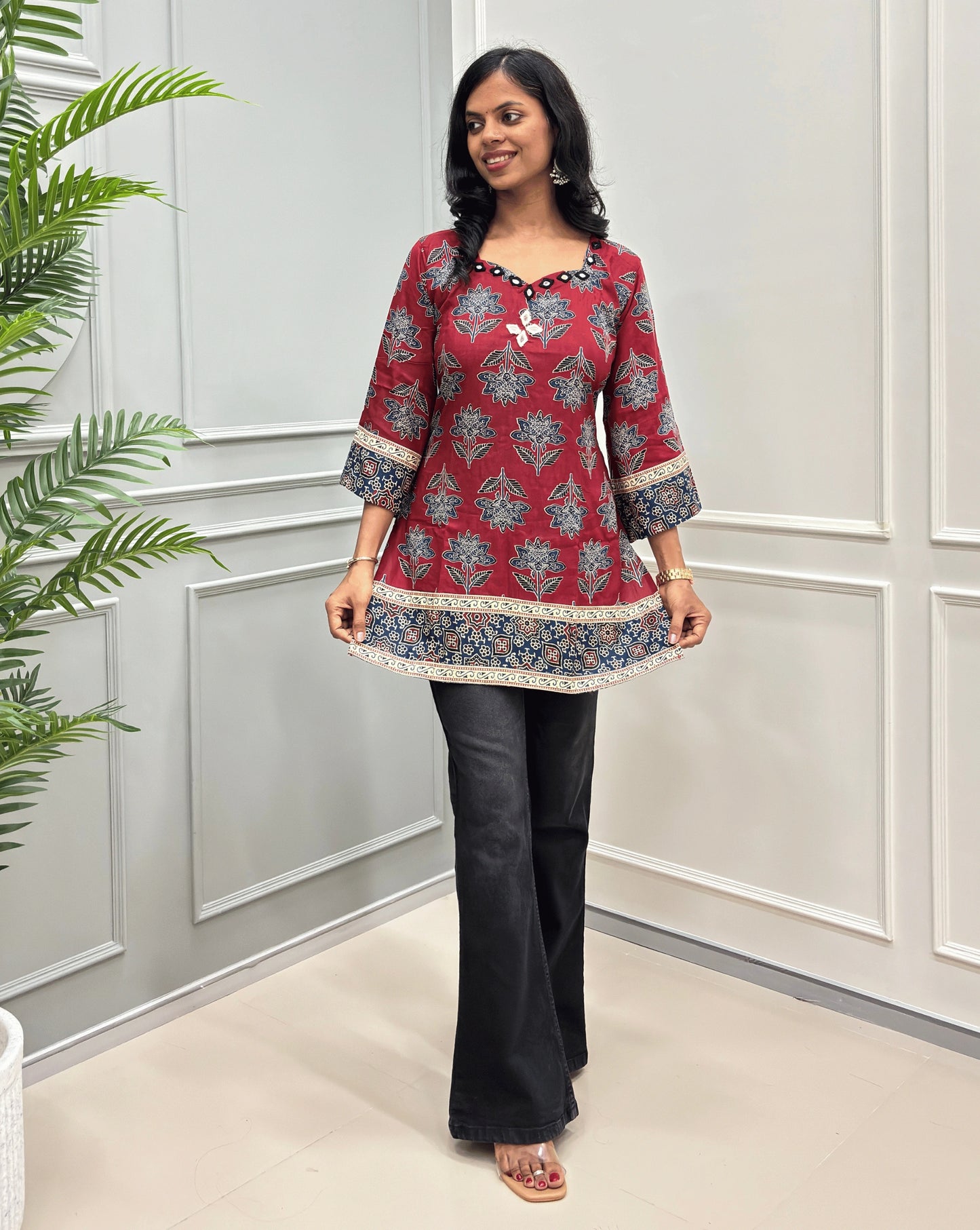 Red Plant based cotton short kurti with blue floral print, paired with black jeans, standing in front of white- grey wall.