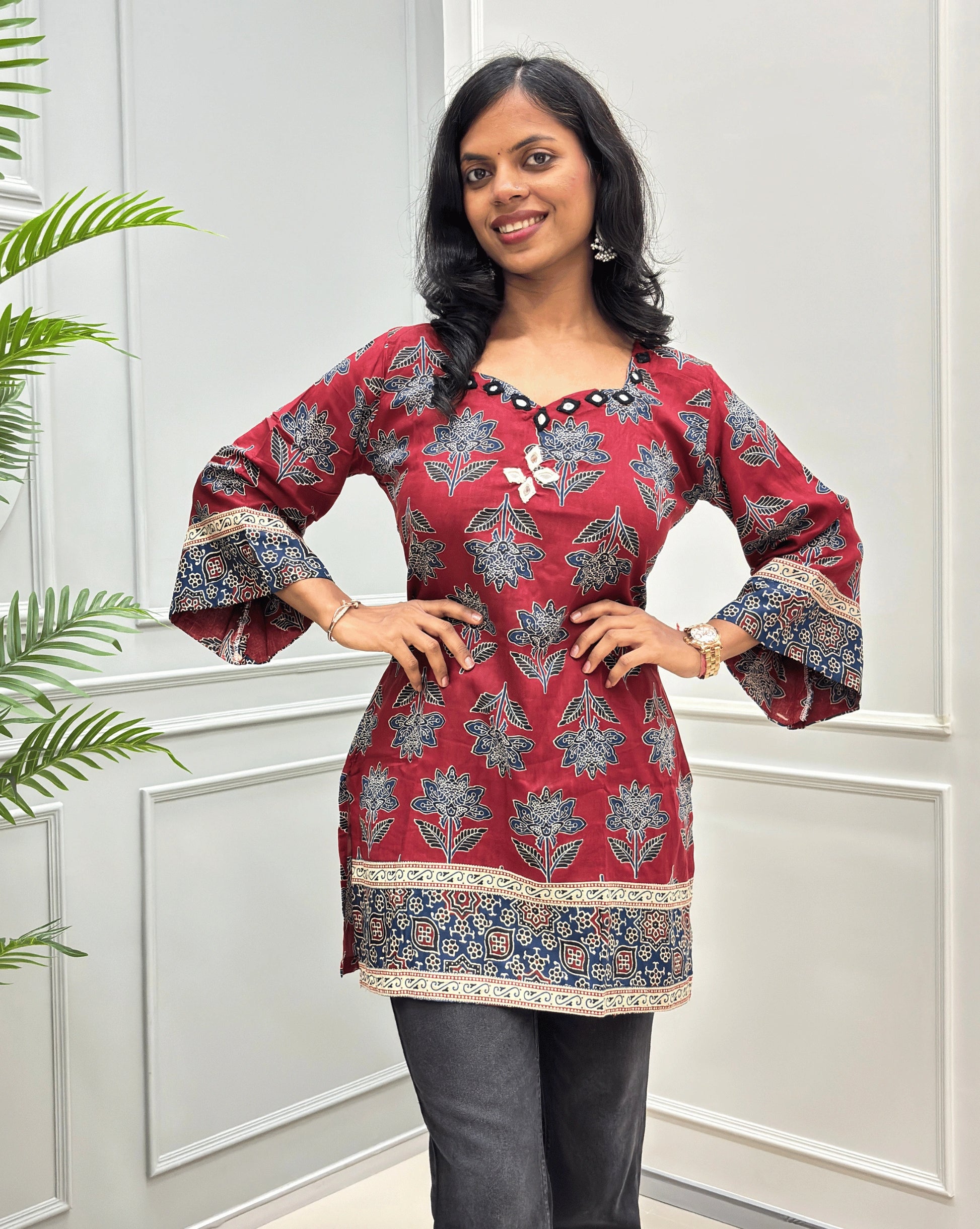 Red women's cotton Ajrakh Short kurti front view with mirror work on neck-line and on chest , blue & beige floral patterns, paired with black flared jeans.