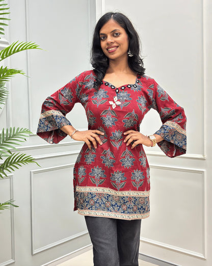 Red women's cotton Ajrakh Short kurti front view with mirror work on neck-line and on chest , blue & beige floral patterns, paired with black flared jeans.