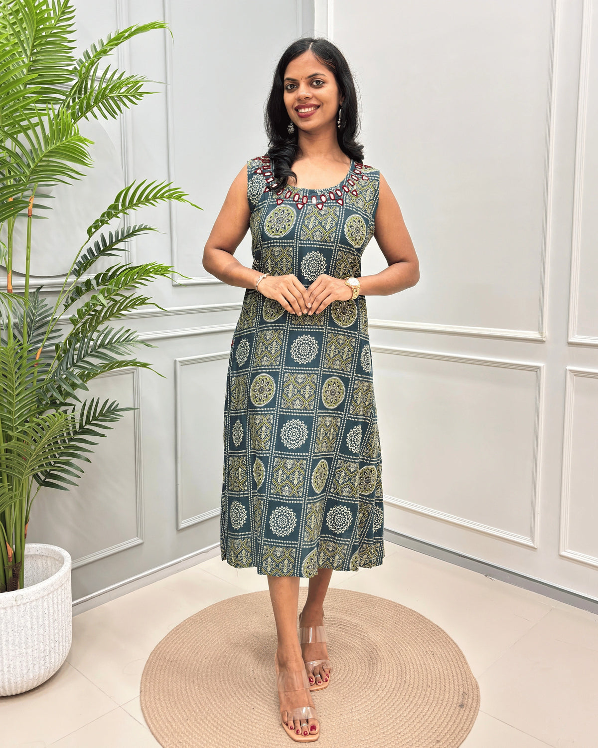 Green Women's Vegetable dyed Plant based Cotton Long-kurti with a green and beige geometric pattern and Hand-Mirror Work on neckline, standing in a decorated room with a potted plant and paneled walls.