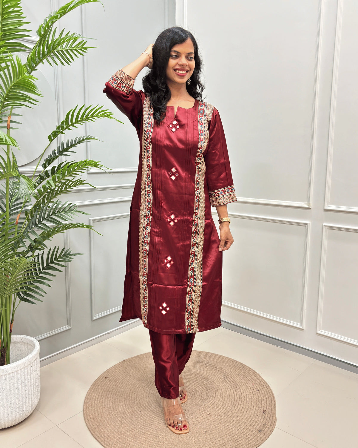 Deep Red Women's Ajrakh Mashru Silk Long Kurti with Multi-color Ajrakh Print on Front & Sleeves, standing on a round woven mat in front of a white paneled wall.