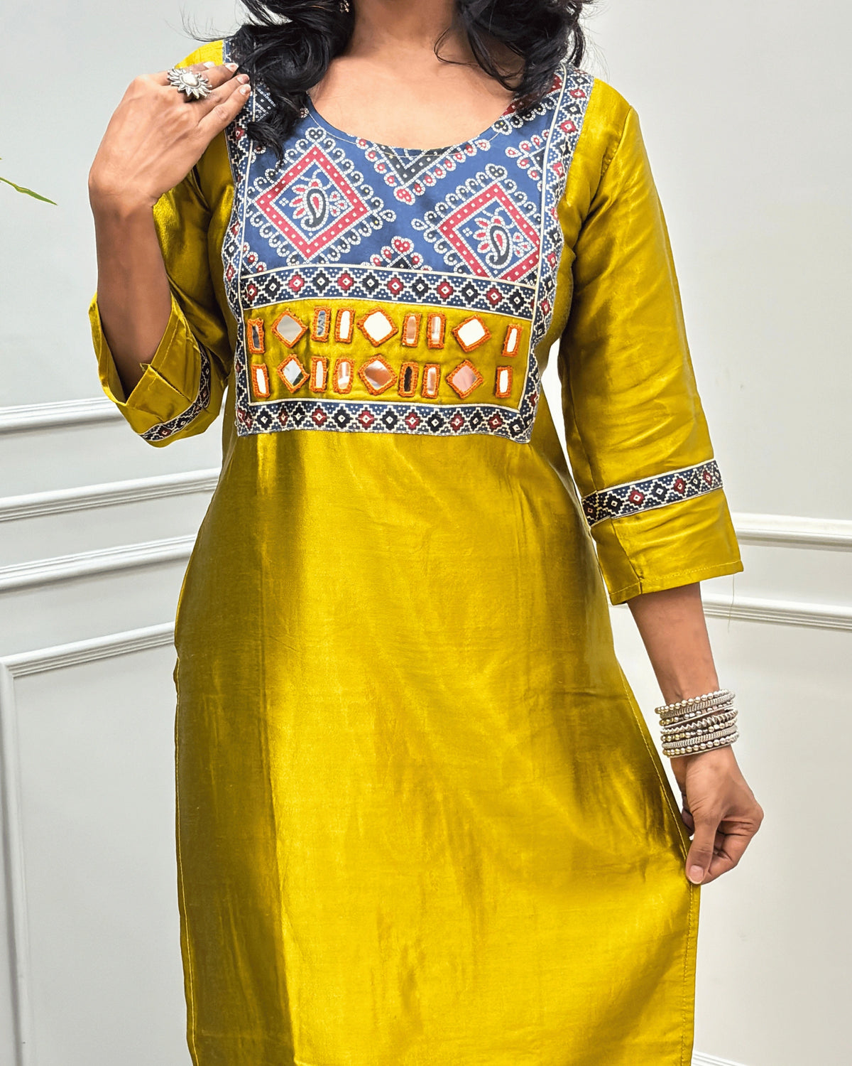 Close-up of Mustard Yellow Ajrakh Mashru Silk Long-kurti with a highly detailed Bandhani embroidered yoke in blue, red, and white, along with ajrakh print border on front & sleeves, posed against a light gray paneled wall.