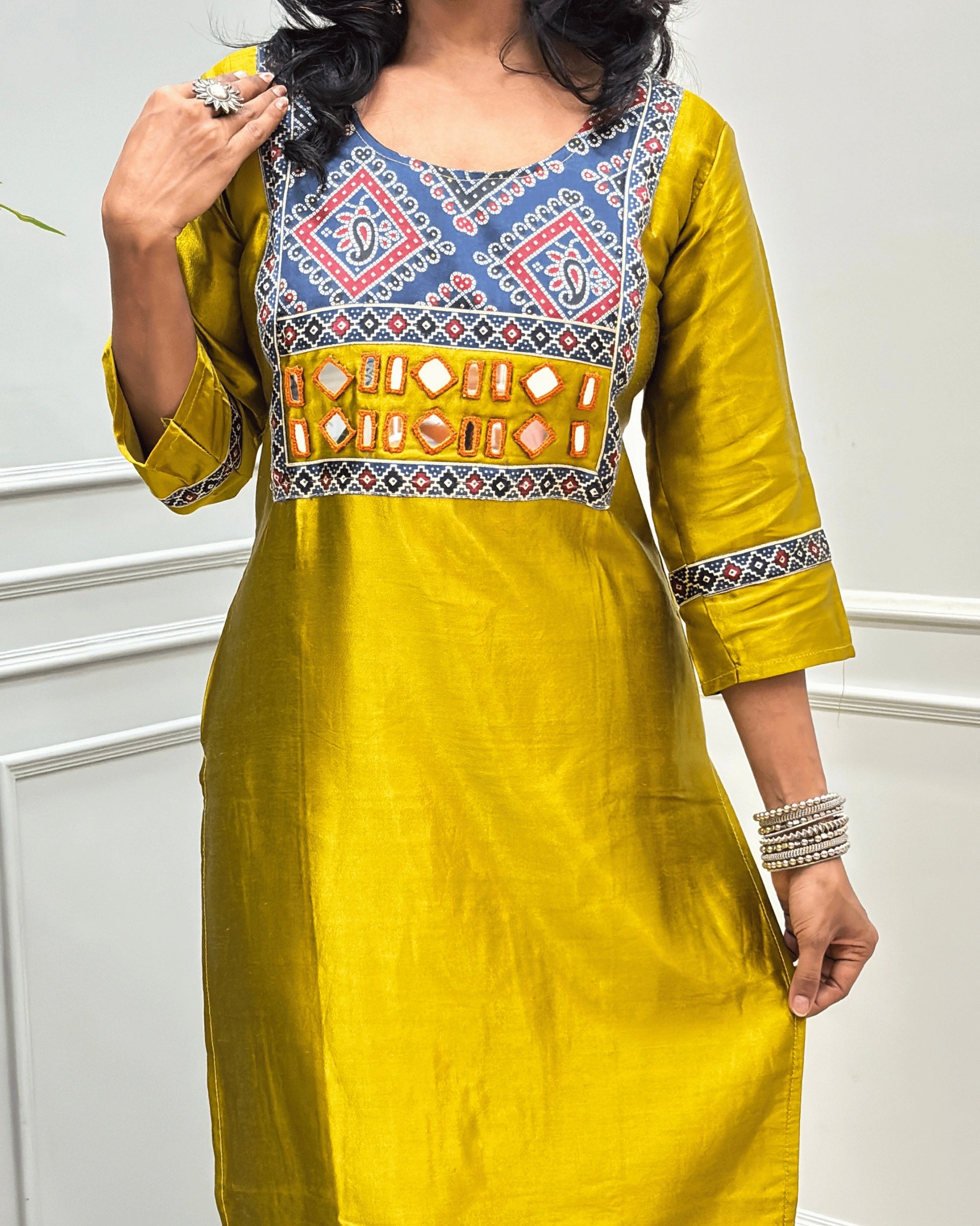 Close-up of Mustard Yellow Ajrakh Mashru Silk Long-kurti with a highly detailed Bandhani embroidered yoke in blue, red, and white, along with ajrakh print border on front & sleeves, posed against a light gray paneled wall.