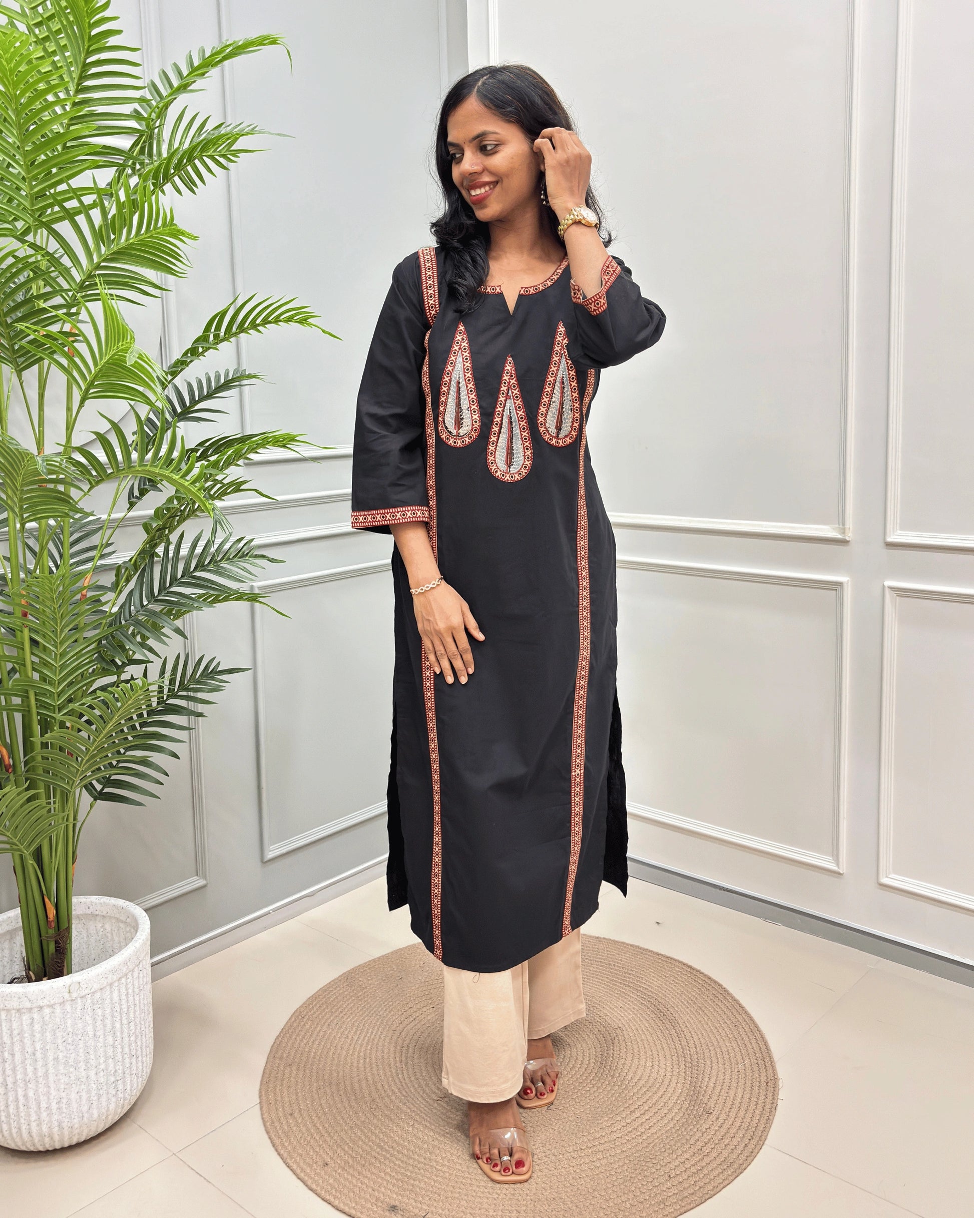 Black Women's Ajrakh Pure Cootn Long-kurti with Beige and Red Ajrakh print with Palm-leaf detailing , paired with beige pants, standing in a room with a potted plant and paneled wall