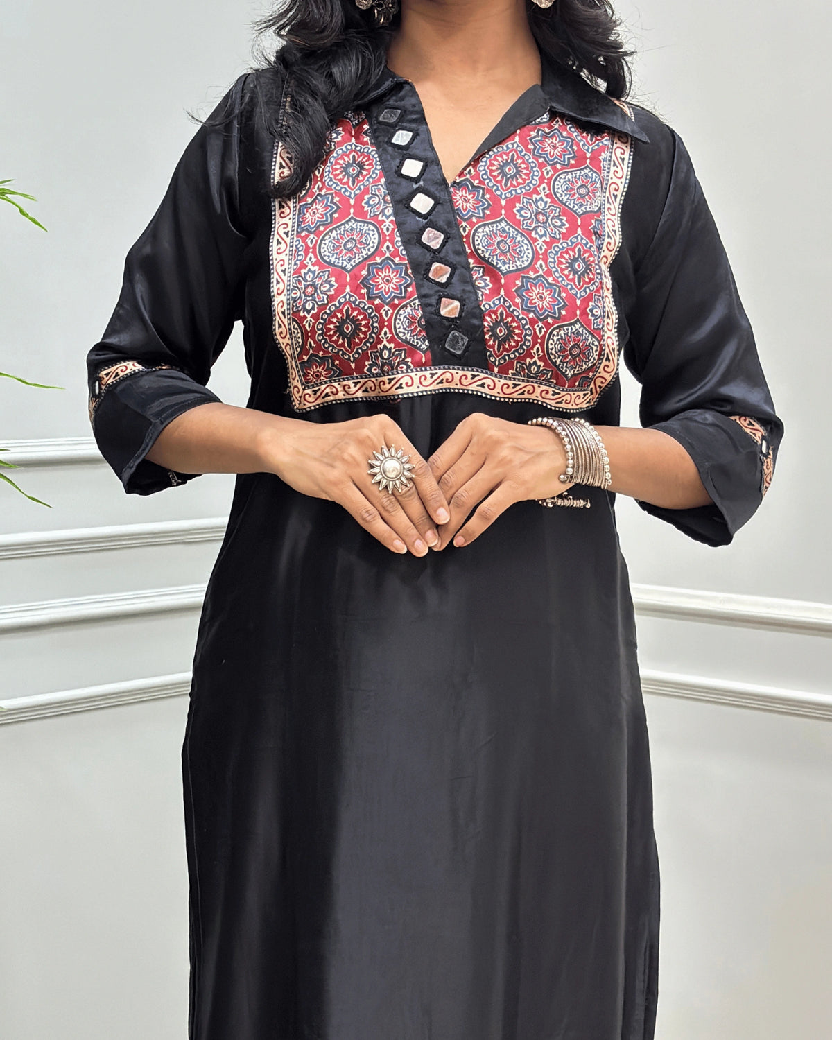 Close-up of Black Ajrakh Mashru Silk Long-kurti with a heavily embroidered Badam-buta yoke and patterned sleeves, Hand-mirror Work on Front & Sleeves, Collar V- neck,  standing against a light gray paneled wall.