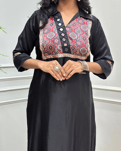 Close-up of Black Ajrakh Mashru Silk Long-kurti with a heavily embroidered Badam-buta yoke and patterned sleeves, Hand-mirror Work on Front & Sleeves, Collar V- neck,  standing against a light gray paneled wall.